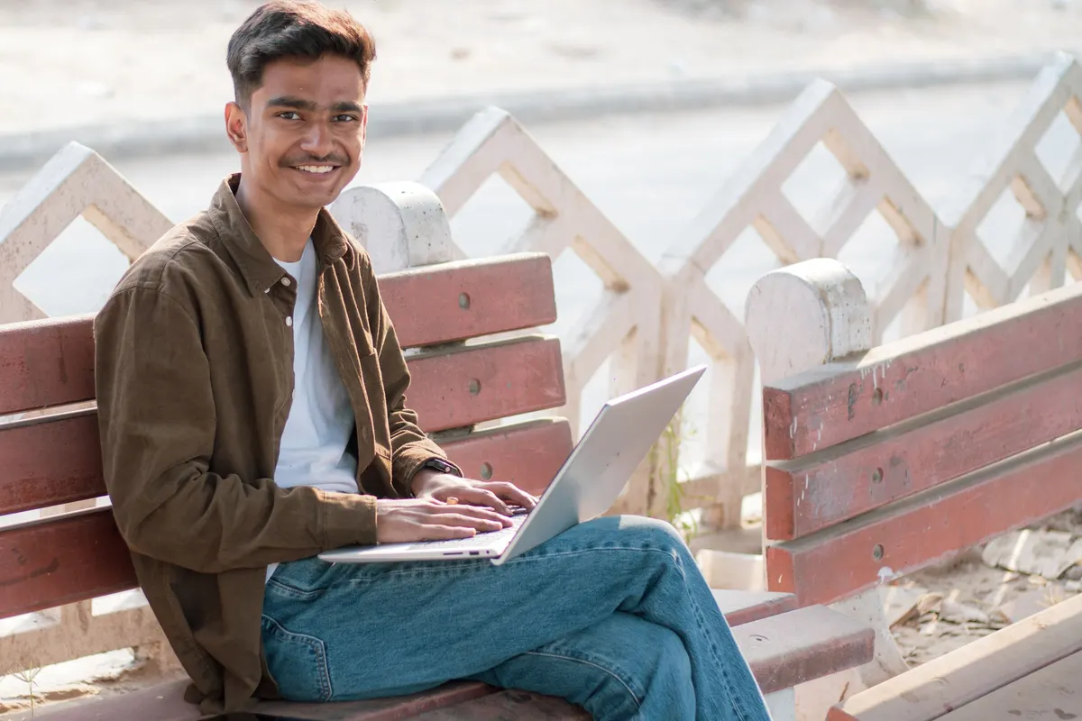 Smiling young man working on a laptop on a park bench